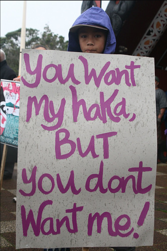 Child holds a large white sign at the May 2009 Hikoi (protest) against the Super City. The signs says "you want my haka, but you don't want me!" in painted pink letters. The child takes up most of the frame, but there is a hint of a Maori meeting house in the background. The child has a determined and solemn expression under their purple raincoat.